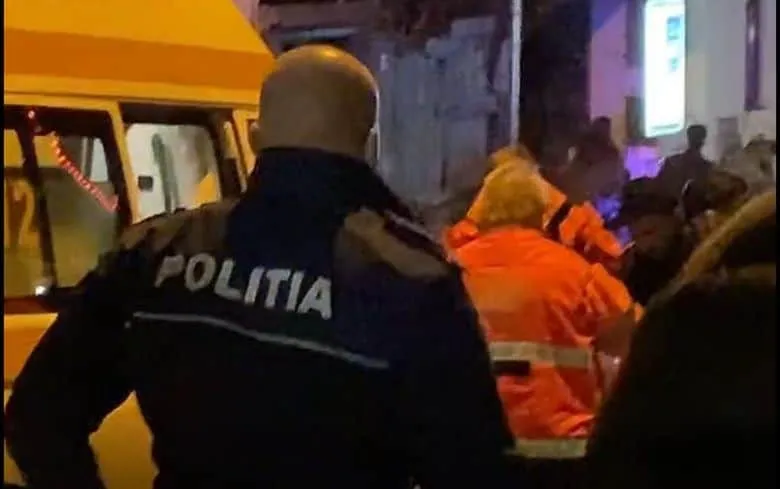 Un agent de poliție cu inscripția POLITIA pe uniformă stă lângă un vehicul galben și persoane îmbrăcate în haine portocalii.