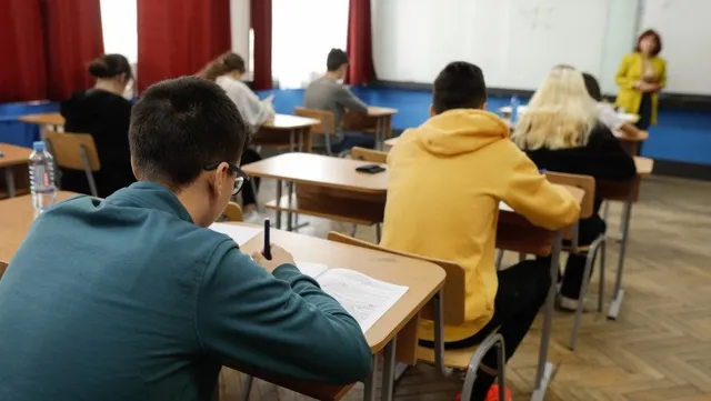 Elevi stau la bănci în interiorul unei săli de clasă, scriind pe hârtii.