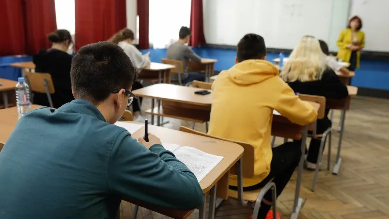 Elevi stau la bănci în interiorul unei săli de clasă, scriind pe hârtii.