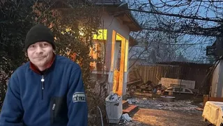 Un bărbat cu căciulă neagră și hanorac albastru stă în fața unei case cu lumină aprinsă în interior și un curte cu vegetație uscată.