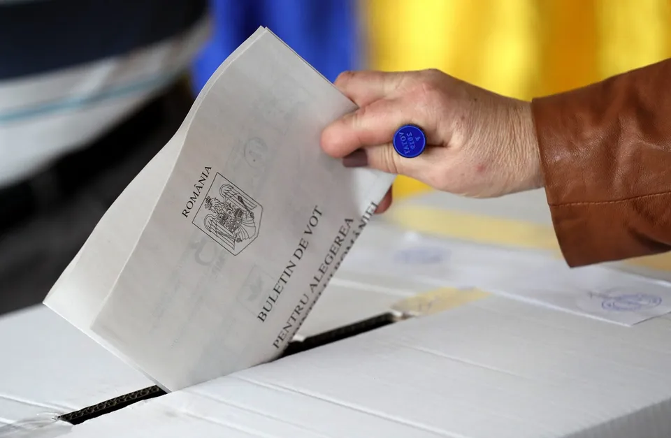 O mână ține un plic alb pe care scrie text și un simbol, introducându-l într-o deschizătură de pe o suprafață albă.