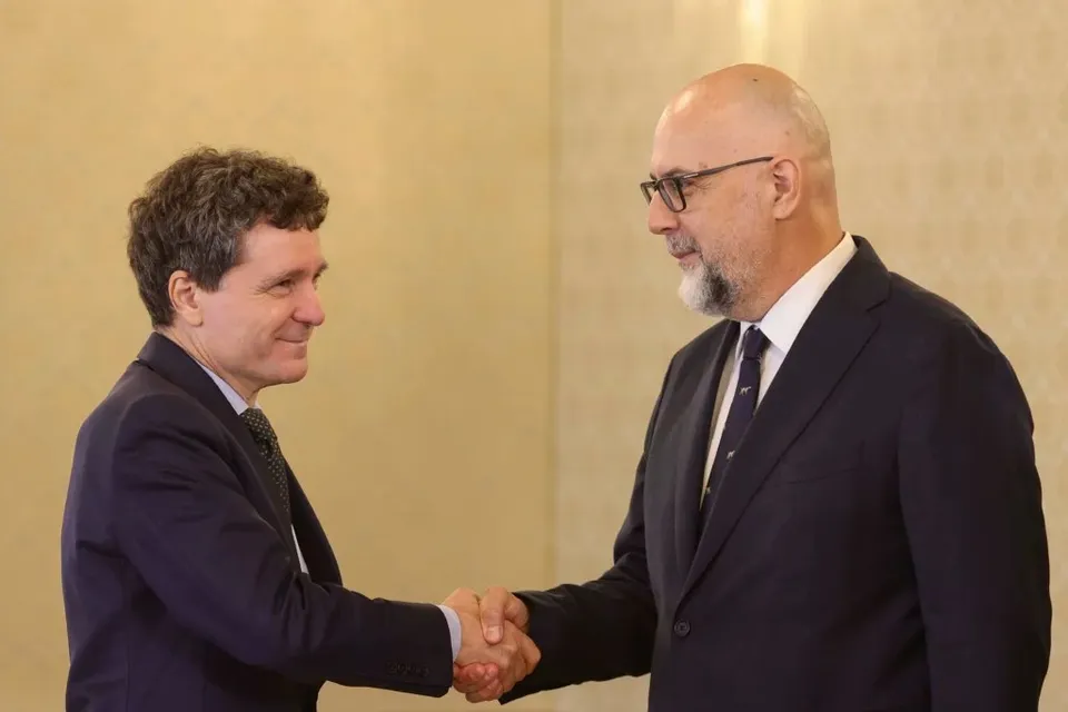 Un bărbat cu părul creț și un bărbat chel cu barbă și ochelari stau față în față și își strâng mâinile.