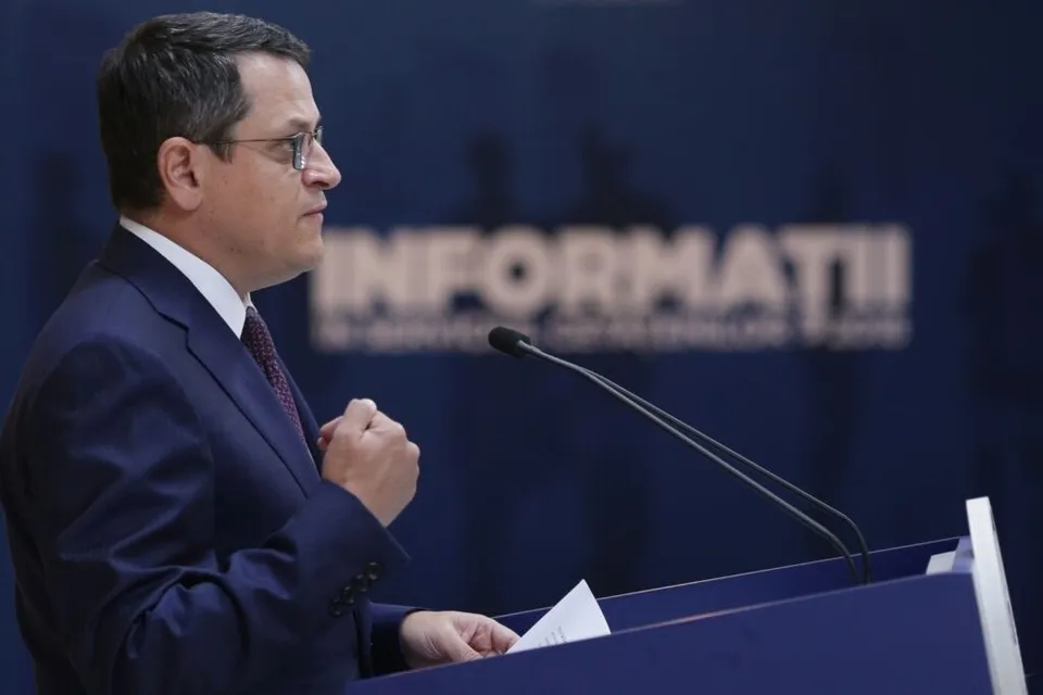 Un bărbat cu ochelari și costum închis la culoare stă în spatele unui podium și privește spre lateral.