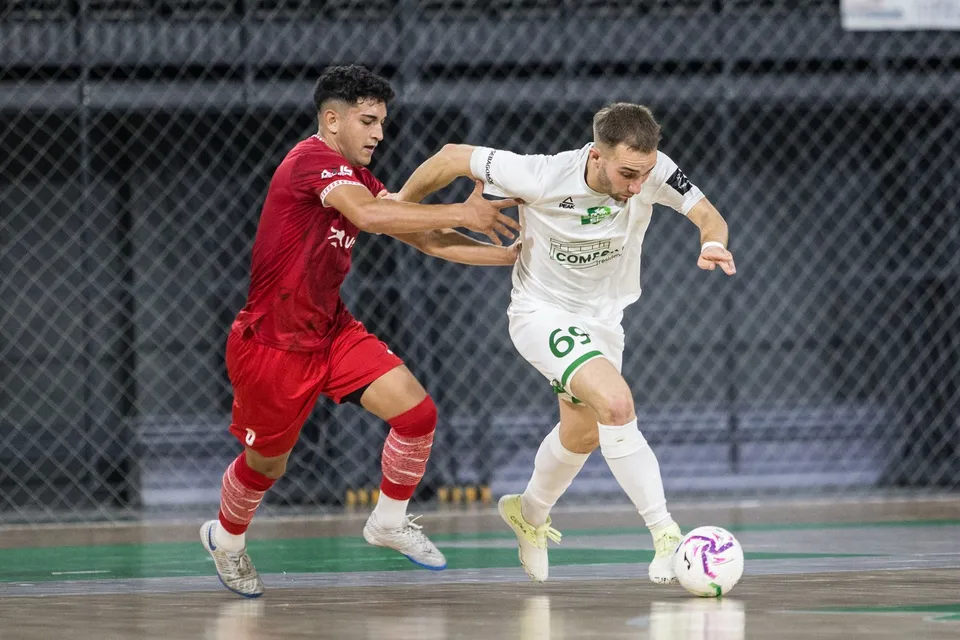Un jucător în echipament alb controlează mingea în timp ce un jucător în echipament roșu încearcă să îi ia posesia pe un teren de futsal.