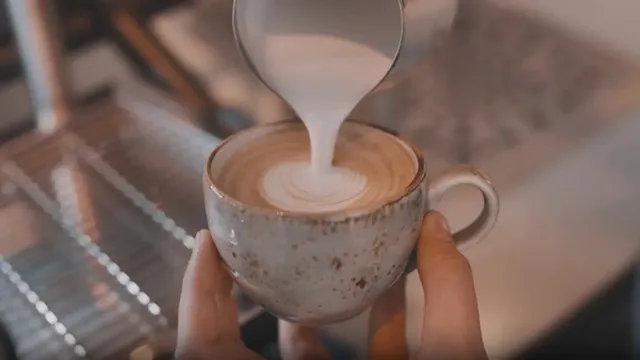 O persoană toarnă lapte spumat dintr-un vas mic într-o cană de ceramică umplută cu cafea.