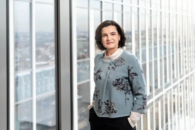 O femeie cu părul scurt și întunecat poartă un sacou albastru cu motive florale și un tricou alb, stând în fața unei ferestre mari.