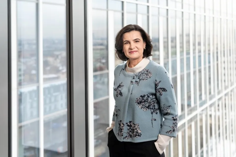 O femeie cu părul scurt și întunecat poartă un sacou albastru cu motive florale și un tricou alb, stând în fața unei ferestre mari.
