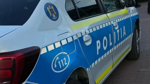 Un autoturism alb cu marcaje albastre și galbene pe care scrie POLITIA și simbolul 112 este parcat pe un drum pavat.