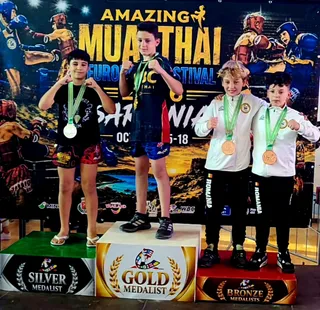Patru copii stau pe podiumuri marcate cu medalii de aur, argint și bronz, purtând medalii și echipamente sportive în fața unui panou colorat.