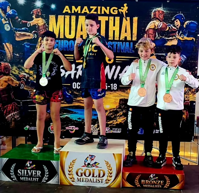 Patru copii stau pe podiumuri marcate cu medalii de aur, argint și bronz, purtând medalii și echipamente sportive în fața unui panou colorat.