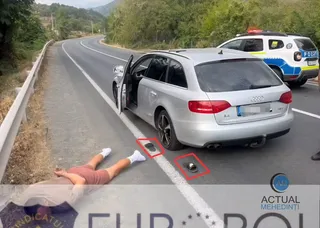 O mașină argintie stă oprită pe marginea unui drum, cu portiera șoferului deschisă, lângă o persoană întinsă pe asfalt în apropierea unei linii albe.