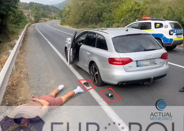 O mașină argintie stă oprită pe marginea unui drum, cu portiera șoferului deschisă, lângă o persoană întinsă pe asfalt în apropierea unei linii albe.