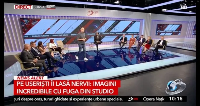 Oamenii stau așezați pe scaune într-un studio de televiziune cu un ecran mare în fundal și un text roșu pe un fundal alb în partea de jos.