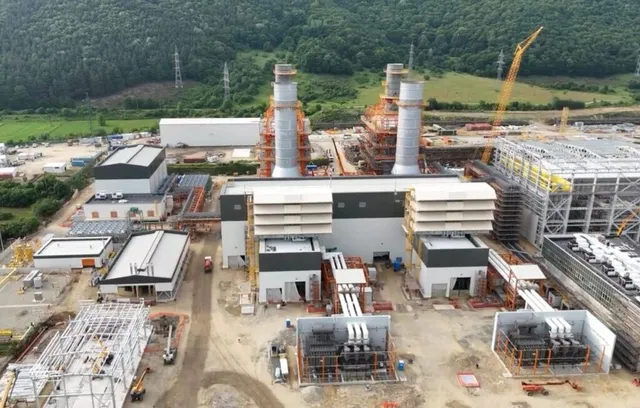 O vedere aeriană a unei centrale electrice în construcție cu două turnuri înalte, structuri metalice și utilaje de șantier pe un teren de pământ.