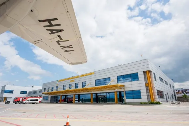 O aripă de avion cu inscripția HA-LXA este poziționată deasupra unei clădiri de terminal de aeroport sub un cer cu nori.