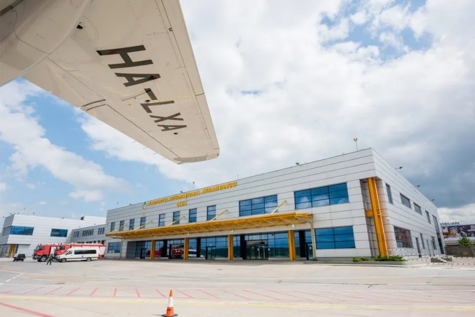 O aripă de avion cu inscripția HA-LXA este poziționată deasupra unei clădiri de terminal de aeroport sub un cer cu nori.