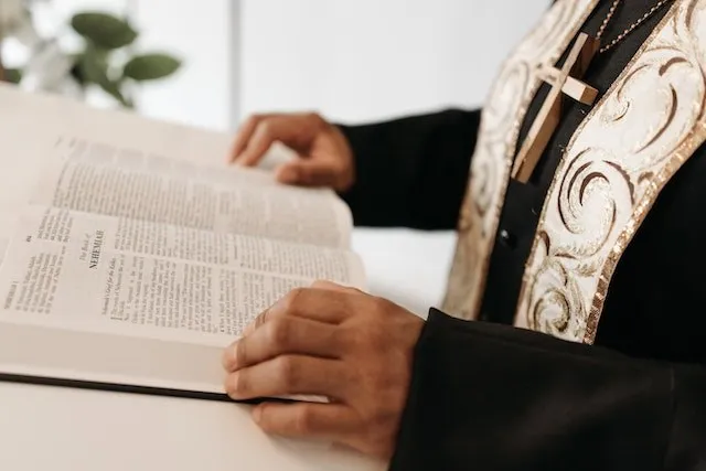 O persoană purtând haine negre cu un ornament decorativ și un crucifix de lemn ține o carte deschisă cu text pe o suprafață albă.