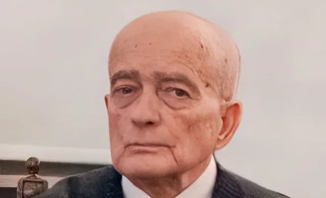 Un bărbat în vârstă cu părul rare și pielea ridată poartă un sacou închis și o cămașă albă pe un fundal deschis.