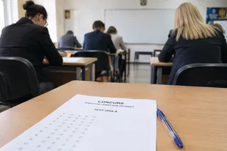 O foaie de test cu cercuri de răspuns și un pix albastru sunt așezate pe o masă de lemn în prim-plan, în fața unor persoane așezate la bănci.