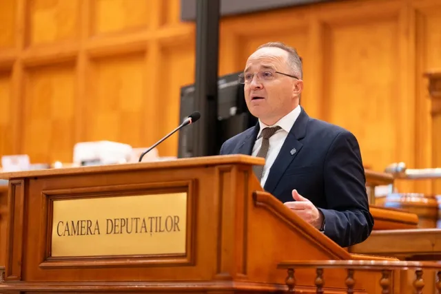 Un bărbat cu ochelari și costum închis stă în spatele unui podium de lemn pe care este scris textul CAMERA DEPUTAȚILOR.