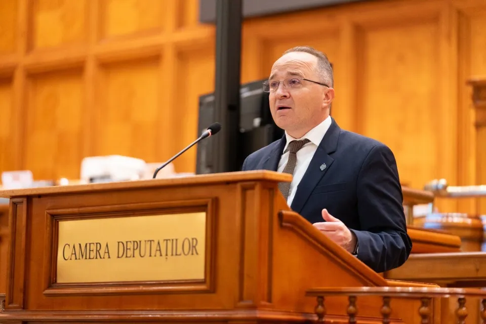 Un bărbat cu ochelari și costum închis stă în spatele unui podium de lemn pe care este scris textul CAMERA DEPUTAȚILOR.
