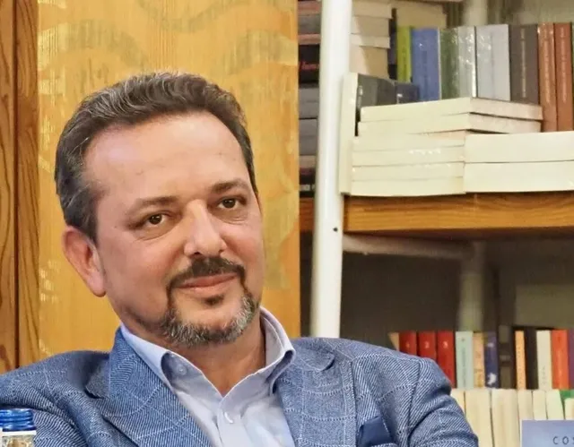 Un bărbat cu barbă și mustață, purtând o sacoșă albastră și o cămașă deschisă la guler, stă în fața unor rafturi cu cărți.