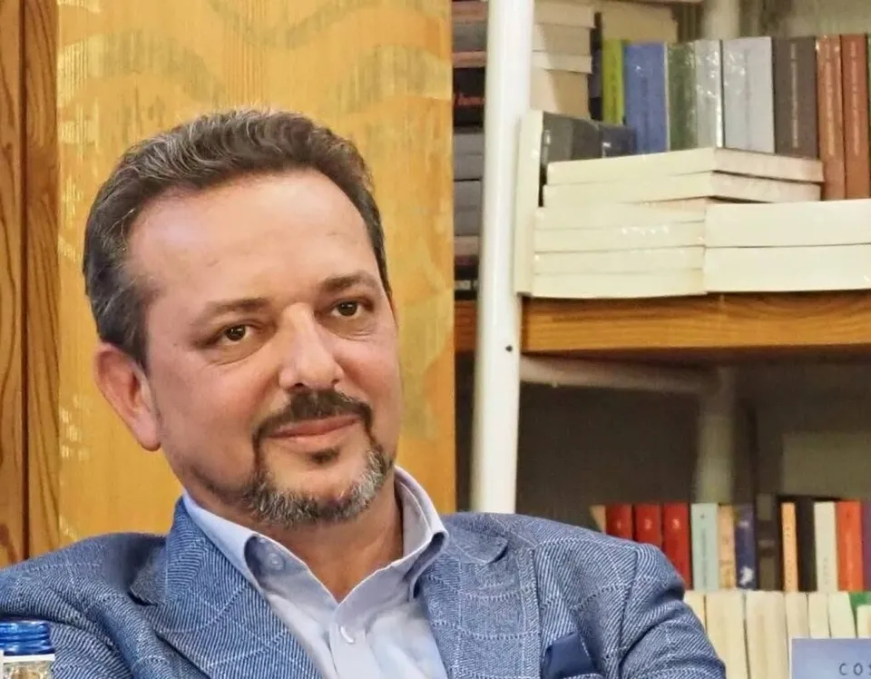 Un bărbat cu barbă și mustață, purtând o sacoșă albastră și o cămașă deschisă la guler, stă în fața unor rafturi cu cărți.