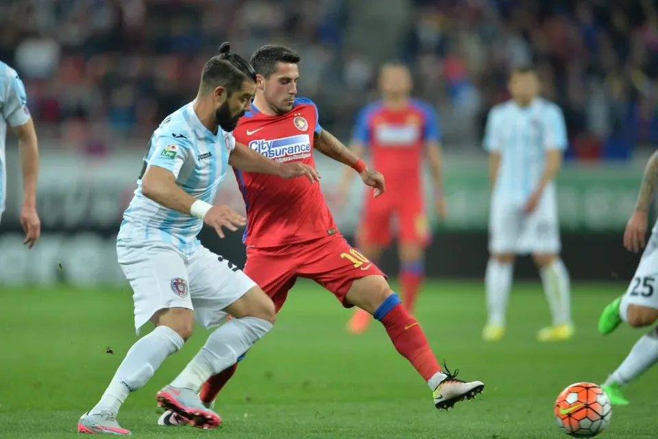 Un jucător în echipament roșu dispută un balon cu un jucător în echipament alb și albastru pe un teren de fotbal.
