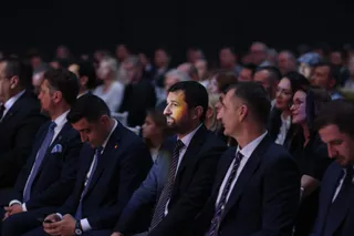 Oamenii stau așezați în rânduri într-o sală întunecată, purtând haine formale precum costume și cravate.