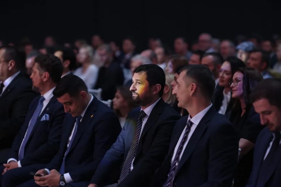 Oamenii stau așezați în rânduri într-o sală întunecată, purtând haine formale precum costume și cravate.