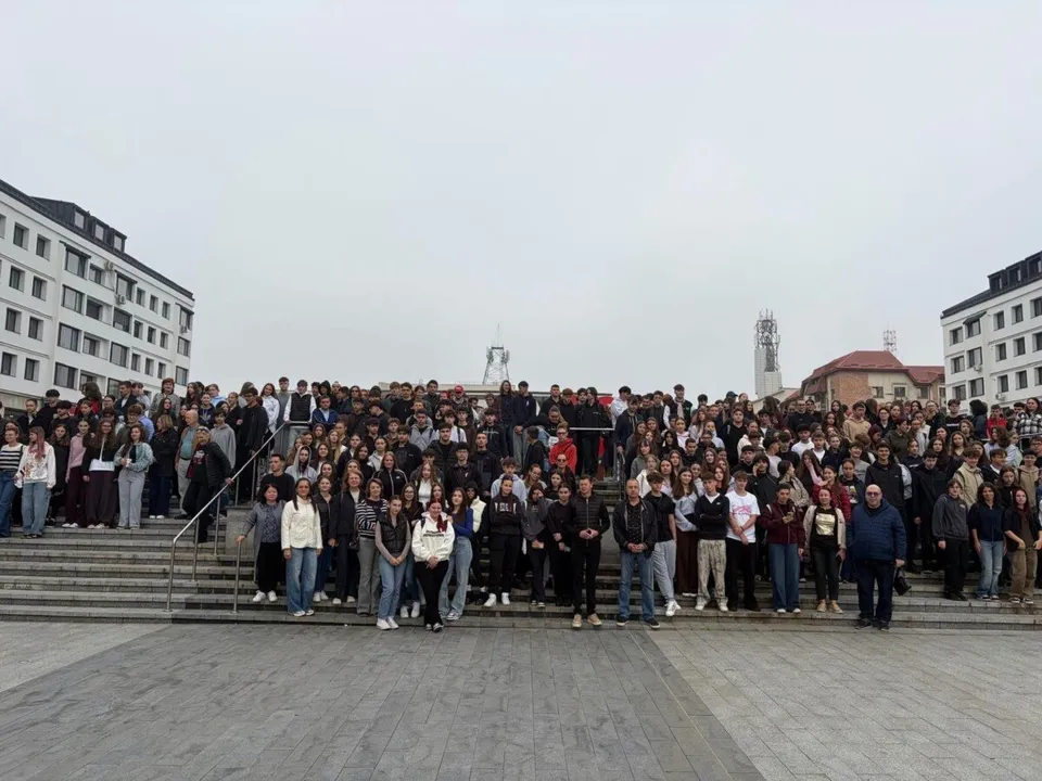 Un grup mare de oameni stă grupate pe scări și pe o platformă pavată sub un cer înnorat.