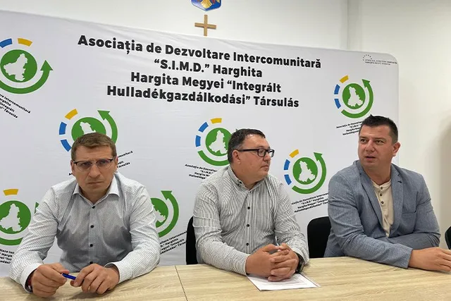 Trei bărbați stau la o masă în fața unui panou alb cu inscripții și logouri verzi și albastre.