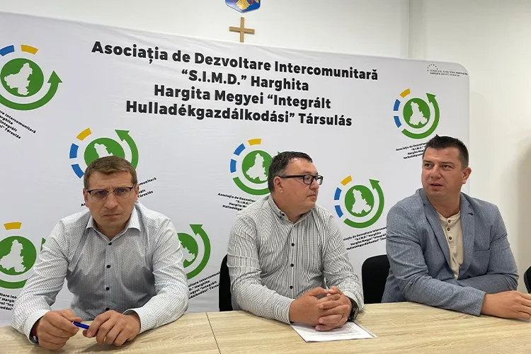 Trei bărbați stau la o masă în fața unui panou alb cu inscripții și logouri verzi și albastre.