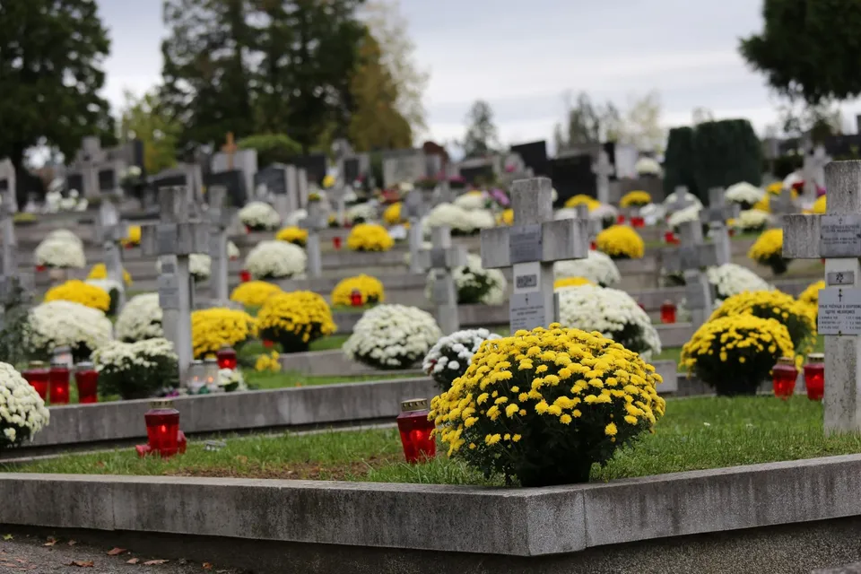 Mai multe morminte cu cruci de piatră sunt decorate cu flori galbene și albe și lumânări mici în interiorul unui cimitir.