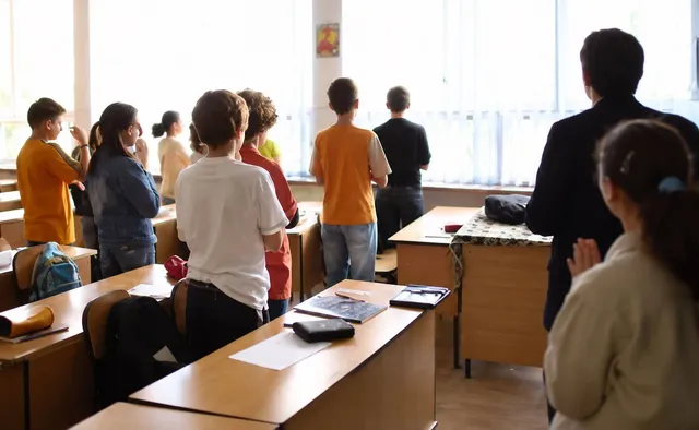 Un grup de elevi stă în picioare în fața unor bănci de școală într-o încăpere luminoasă cu ferestre mari.
