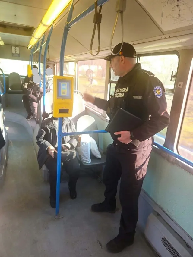 Un bărbat în uniformă neagră cu inscripții și un aparat portabil stau în interiorul unui vehicul public lângă pasageri așezați pe scaune.