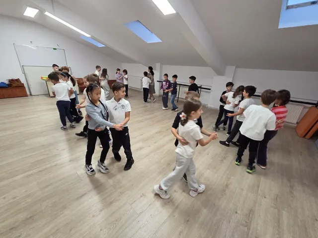 Elevi în uniformă școlară sau vestimentație casual danând în perechi pe un parchet de lemn într-o sală cu pereți albi și ferestre în tavan.