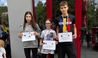 Trei tineri stau în picioare purtând medalii și ținând certificate de participare în fața unei clădiri cu cadre roșii.