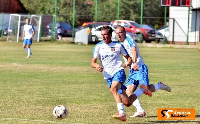 Un jucător de fotbal în echipament alb și albastru conduce mingea pe un teren de iarbă, fiind urmărit de un alt jucător în echipament similar.