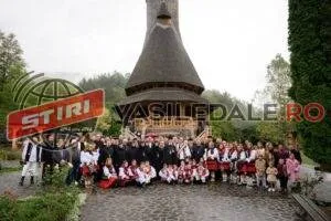 Un grup mare de persoane îmbrăcate în costume tradiționale stă poziționat în fața unei biserici din lemn cu acoperiș înalt și ascuțit.