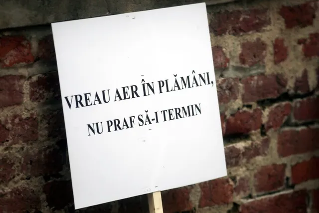 Un panou alb cu text negru pe care scrie „VREAU AER ÎN PLĂMÂNI, NU PRAF SĂ-I TERMIN” este fixat pe un zid de cărămidă roșie.