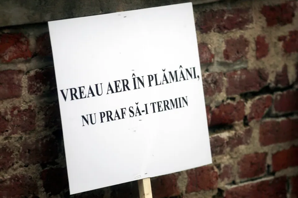 Un panou alb cu text negru pe care scrie „VREAU AER ÎN PLĂMÂNI, NU PRAF SĂ-I TERMIN” este fixat pe un zid de cărămidă roșie.