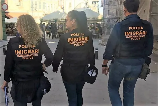Trei persoane purtând jachete negre cu inscripția IMMIGRATION POLICE merg pe o stradă lângă mese de cafea sub umbrele de soare.