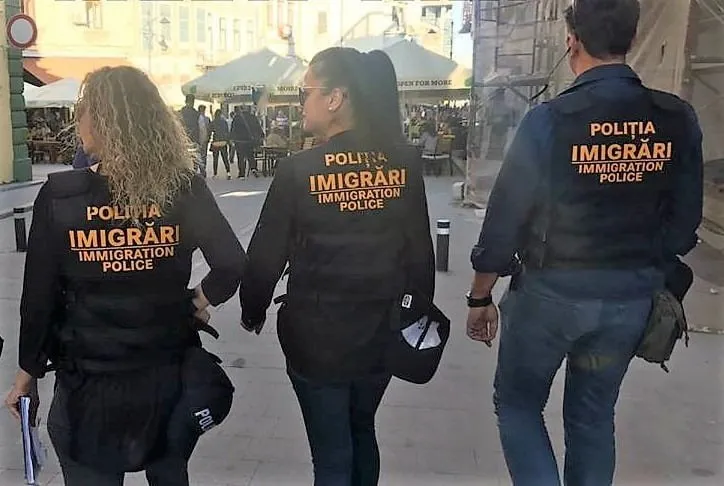 Trei persoane purtând jachete negre cu inscripția IMMIGRATION POLICE merg pe o stradă lângă mese de cafea sub umbrele de soare.