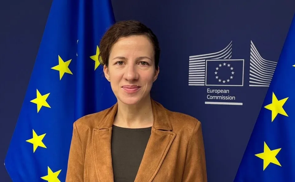 O femeie cu părul scurt și o jachetă maro stă în fața unui fundal albastru cu stele galbene și logo-ul Comisiei Europene.