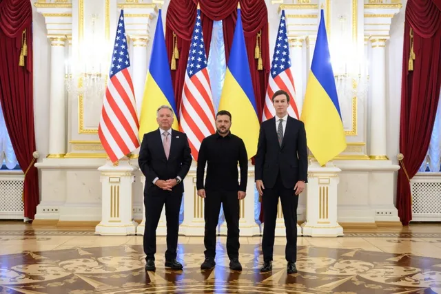 Trei bărbați stau în picioare în fața unor steaguri americane și ucrainene într-o încăpere decorată cu coloane și draperii roșii.