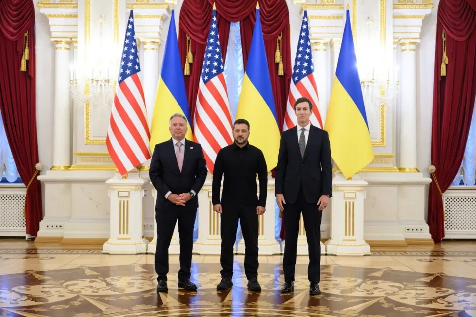 Trei bărbați stau în picioare în fața unor steaguri americane și ucrainene într-o încăpere decorată cu coloane și draperii roșii.
