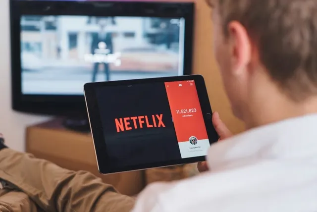 Un bărbat cu cămașă albă ține o tabletă pe care se afișează logo-ul Netflix și un număr pe un fundal roșu.
