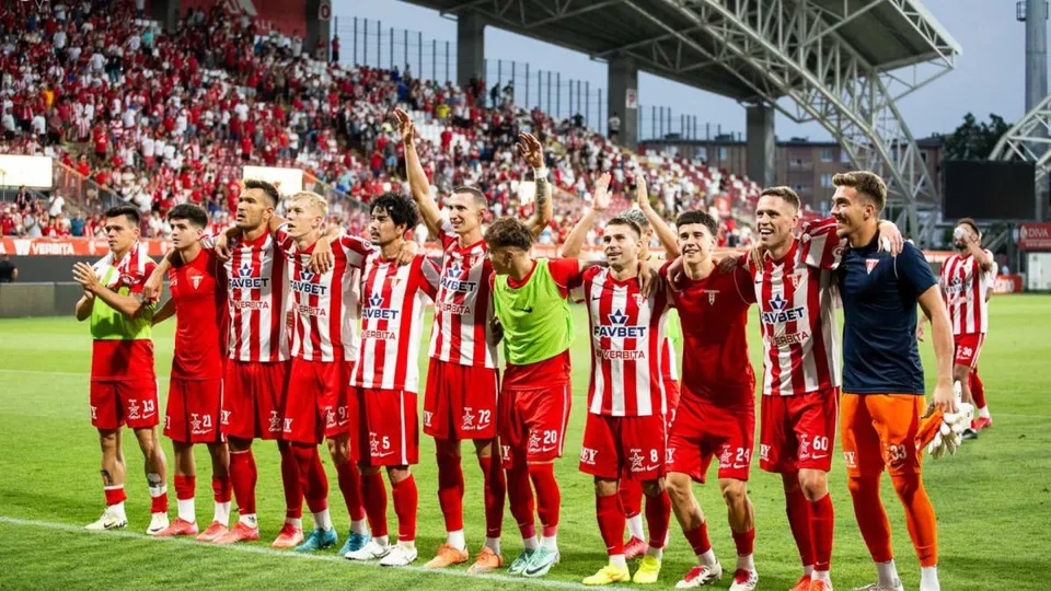 O echipă de fotbaliști în echipament roșu și alb sărbătorește pe un teren de fotbal în fața unor tribune pline de spectatori.