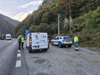 Unii oameni în veste fluorescente stau lângă un van alb și o mașină de poliție pe marginea unui drum asfaltat lângă un deal împădurit.